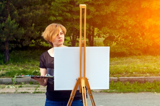 Artist  young woman paints on a canvas an urban landscape of a summer of oil paints in city park