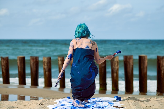 Artistic blue-haired woman performance artist in dark blue dress smeared with blue gouache painting and dancing on beach, back view. Body painting, body art concept, expressive performance art outdoor