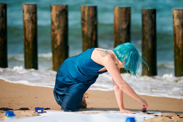 Artistic blue-haired woman performance artist in dark blue dress smeared with blue gouache painting with wide strokes on large canvas on beach. Expressive performance art, contemporary art outdoor