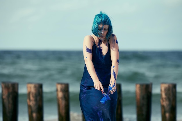 Artistic blue-haired woman performance artist in dark blue dress smeared with indigo gouache painting on her body with brushes on beach. Creative body painting, body art concept, outdoor performance