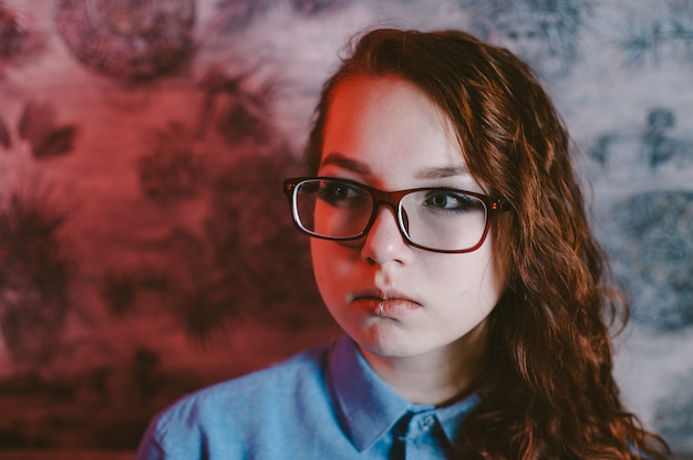 Artistic portrait of a beautiful red-haired girl wearing red glasses and a blue shirt. Piercing in the lip.