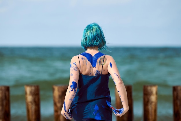 Artistieke blauwharige vrouw performance artiest in donkerblauwe jurk besmeurd met indigo gouache schilderij op haar lichaam met borstels op strand, achteraanzicht. Creatief bodypainten, body art concept, buiten