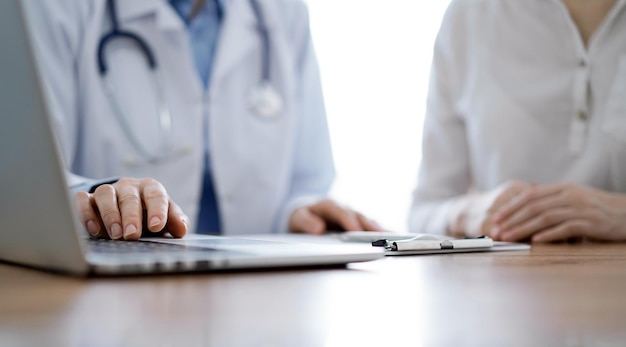 Arts en patiënt bespreken iets terwijl ze een laptop gebruiken en naast elkaar zitten aan het houten bureau in de kliniek. Geneeskunde concept