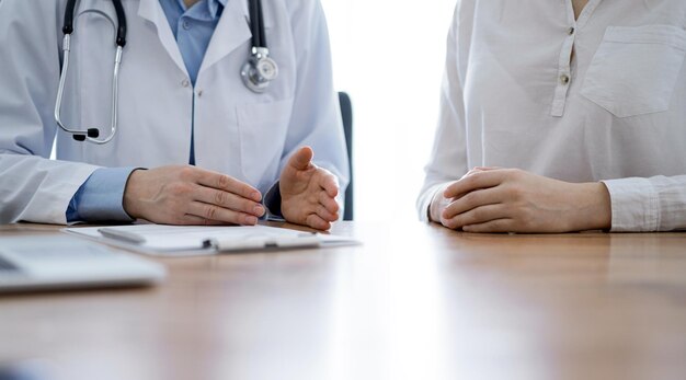 Arts en patiënt bespreken iets terwijl ze naast elkaar aan het houten bureau in de kliniek zitten, close-up van handen. Geneeskunde concept.