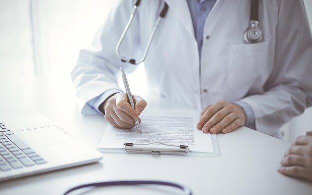 Arts en patiënt zitten aan tafel in de kliniek. De focus ligt op de handen van vrouwelijke artsen die het medicatiegeschiedenisformulier of de checklist invullen, van dichtbij. Geneeskundeconcept.