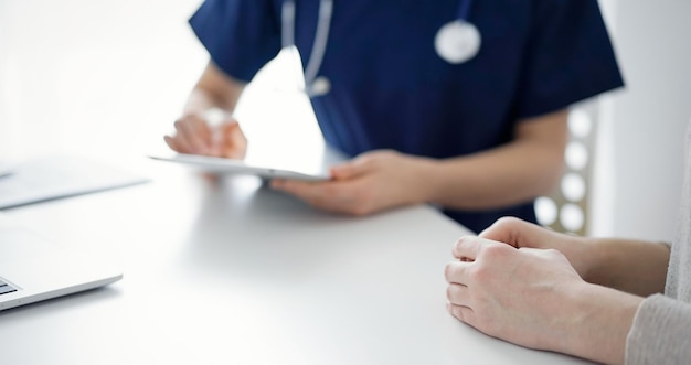 Arts en patiënt zitten aan tafel in de kliniek terwijl ze iets bespreken en een tabletcomputer gebruiken. De focus ligt op de handen van vrouwelijke patiënten, van dichtbij. Geneeskundeconcept.