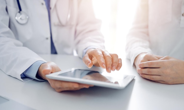 Arts en patiënt zitten naast elkaar aan het bureau in de kliniek. De focus ligt op de handen van de vrouwelijke arts die naar het touchpad van de tabletcomputer wijzen, close-up. Geneeskunde concept.