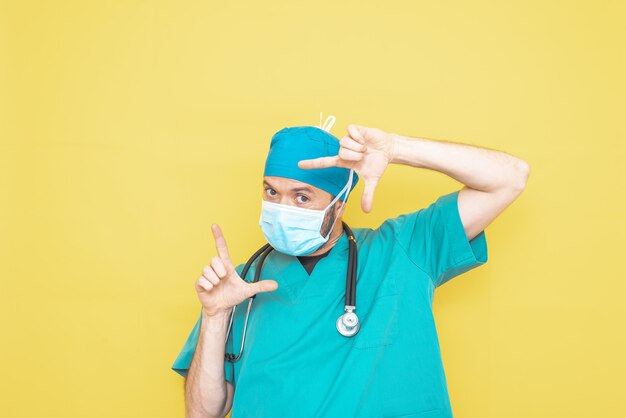 Arts gekleed als chirurg in het groen met een stethoscoop en masker op een gele achtergrond met een bezorgde uitdrukking.