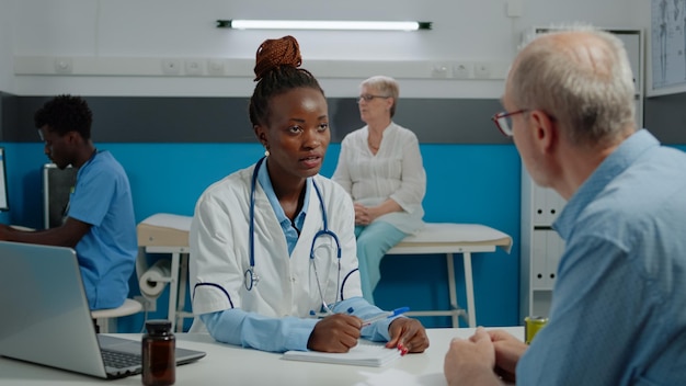 Arts in gesprek met senior zieke patiënt in zorgkantoor. Dokter die controleregistratiepapieren geeft aan medische assistent op de achtergrond. Oude persoon met ziekte in gesprek met specialist