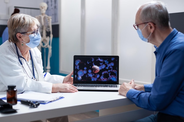 Arts legt coronavirusbacteriën op laptop uit aan senior man, met gezichtsmasker in medisch kantoor. vrouwelijke arts die een visuele weergave van de viruspandemie toont bij controlebezoek met de patiënt