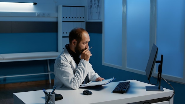 Arts-medic staat aan bureautafel en kijkt naar gegevensexpertise op de computer in vergelijking met medische papieren terwijl hij 's avonds laat in het ziekenhuiskantoor werkt. Medicijnservice en concept