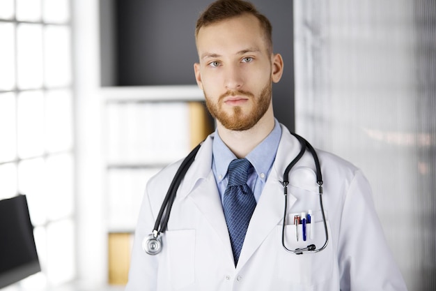 Foto arts met een rode baard staat recht in de kliniek in de buurt van zijn werkplek. portret van arts. geneeskunde, gezondheidszorg.