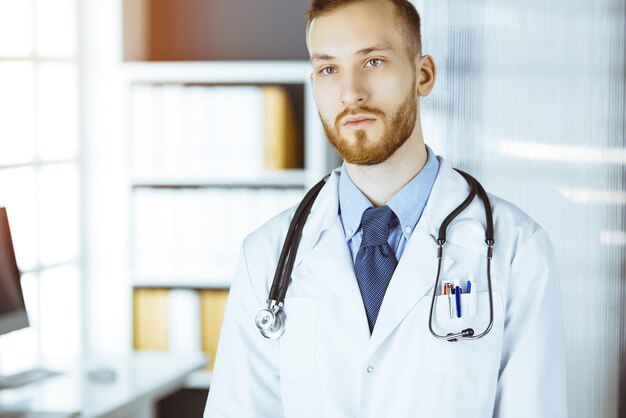 Arts met rode baard die rechtop staat in een zonnige kliniek in de buurt van zijn werkplek Portret van arts Geneeskundeconcept