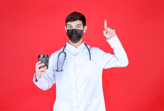 Arts met stethoscoop in zwart masker die een zwarte afhaalkoffiekop houdt en denkt.