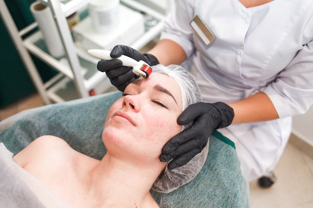 Arts-schoonheidsspecialist maakt een gezichtsmassage met een dermo-roller. Vrouw in schoonheidssalon tijdens mesotherapie procedure met mesoscooter