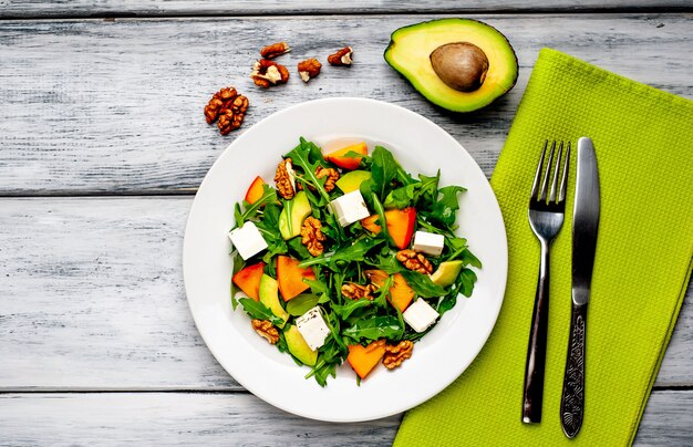 나무 테이블에 흰 접시에 Arugula 샐러드, 감, 호두, 아보카도, 죽은 태아의 치즈