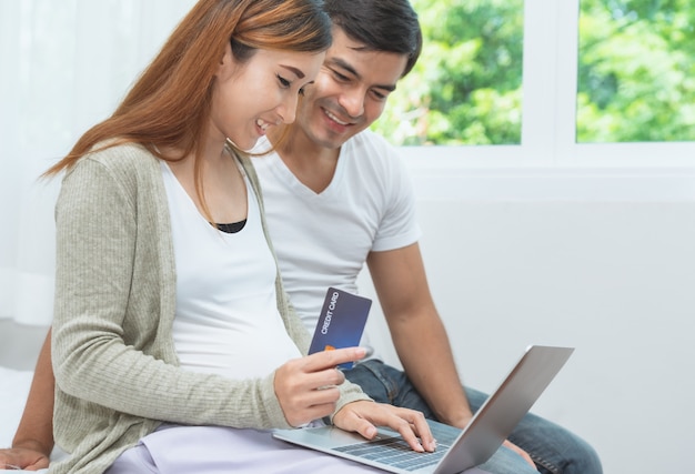 Asain pregnant woman and her husband shopping online on laptop computer and payment by credit card at home