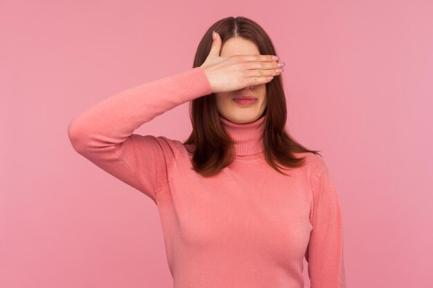 Ashamed confused woman with brown hair closing eyes with hand hiding ignoring dont want to see Indoor studio shot isolated on pink background