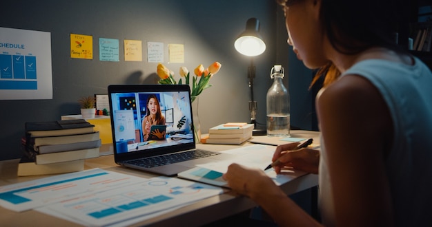 Asia businesswoman using laptop talk to colleagues about plan in video call meeting at living room at home. Working from house overload at night, remotely, social distance, quarantine for coronavirus.