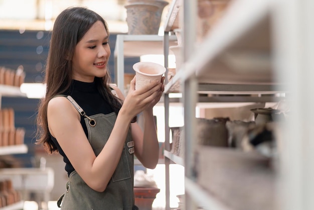 앞치마를 든 아시아 여성 도예가는 자신의 새로운 컬렉션을 선보인 것을 자랑스럽게 생각하며 웃고 있는 오븐아시아 여성 중소기업 세라믹 워크샵에서 굽기를 기다리는 선반에 점토 조각 제품을 정렬합니다.