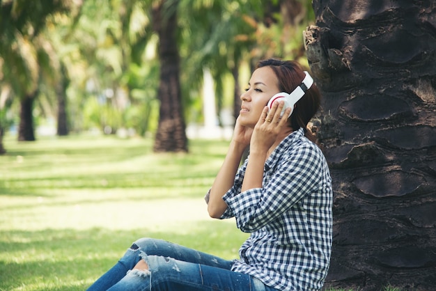 La donna dell'asia gode dell'ascolto della musica in linea sotto l'albero al parco pubblico. rilassarsi tecnologia internet del concetto di cose