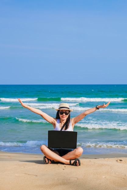 Foto donna dell'asia che si siede sulla spiaggia e che per mezzo del computer portatile per il suo lavoro e controllando affare durante la sua vacanza.