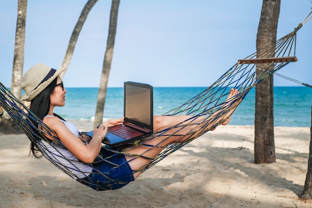 asia woman sitting on the hammock near the beach and using laptop for her work and checking business during her vacation.