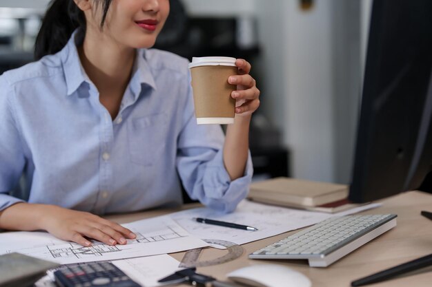 Asian architect engineer women reading architecture information plan on computer and drinking hot chocolate while working to checking about drawing of building and construction plan on blueprint