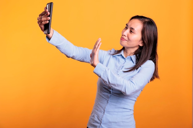 Asian attractive woman greeting remote people during mobile phone videocall meeting, enjoying conversation in studio over yellow background. confident adult having fun with friend during call