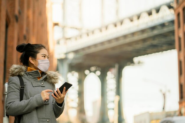 Asian attractive Woman traveller wearing a face pollution mask to protect herself from the coronavirus with background of Down Under the Manhattan Bridge Overpass near the Manhattan Bridge Newyork US