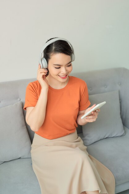 Asian attractive woman wearing headphones, using a mobile phone and sitting on sofa