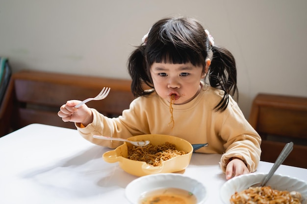 ダイニングテーブルのキッチンでおいしい麺を食べるアジアの赤ちゃん。幸せなアジアの女の赤ちゃんは、ダイニングテーブルで自分で食べることを練習します。赤ちゃんのライフスタイルのコンセプト