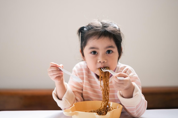 La bambina asiatica si diverte felice usando il cucchiaio e la forchetta delle posate mangiando una deliziosa tagliatella in cucina sul tavolo da pranzo la bambina asiatica felice si esercita a mangiare da sola sul tavolo da pranzo concetto di alimenti per bambini