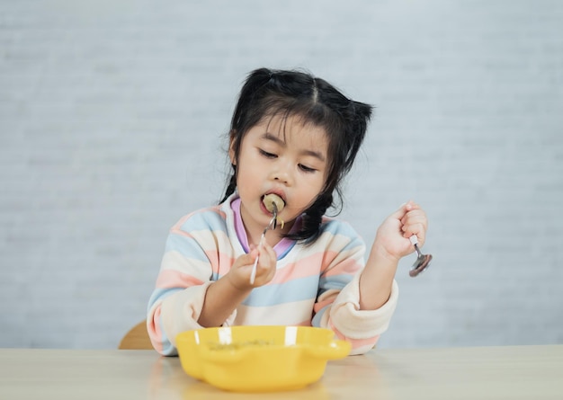 Азиатская девочка счастлива, используя ложку и вилку для столовых приборов, поедая вкусную лапшу и фрикадельки на кухне на обеденном столе Счастливая азиатская девочка практикуется есть сама на обеденном столе Концепция детского питания