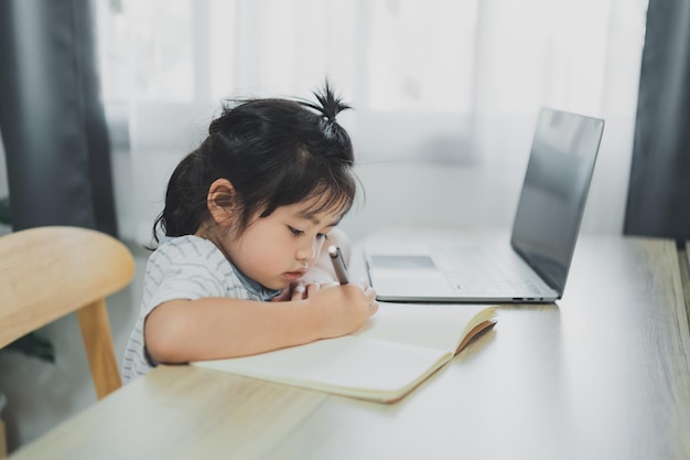 Asian baby girl wearing a blue striped shirt use laptop and\
write notes in notebook to study online on wood table desk in\
living room at home education learning online from home\
concept