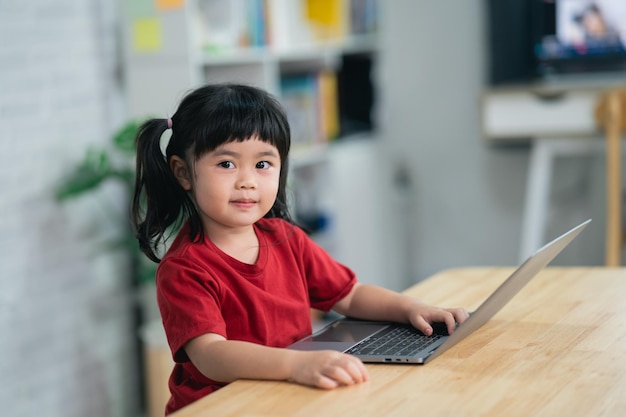 Asian baby girl wearing red tshirt smile and laugh while use laptop and study online on wood table desk in living room at home Education learning online from home concept