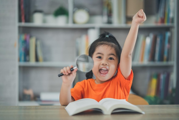 Ragazza asiatica del bambino del bambino che tiene la lente d'ingrandimento e alza la mano leggendo il libro educazione bambini e concetto di scuola ragazza studentessa sorridente felice che impara studiando il concetto di sviluppo dell'istruzione