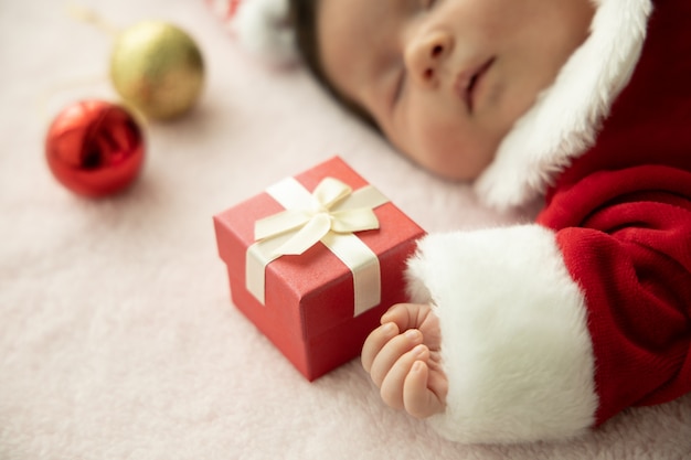 Bambino asiatico neonato sull'uniforme del babbo natale che dorme con il presente della scatola rossa e il cappello rosso