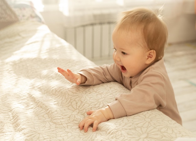 An Asian baby stands in the bedroom leaning on the bed and trying to take her first steps