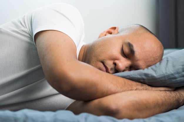 Азиатский лысый мужчина лет 30 в белой футболке спит.