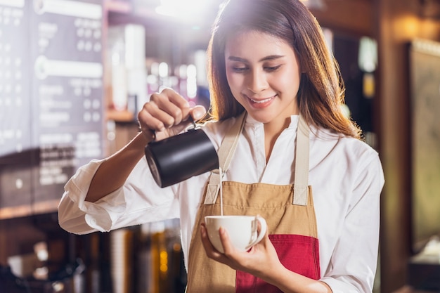 顧客のためにラテまたはカプチーノとエスプレッソであるコーヒーのカップにミルクを注ぐアジアのバリスタ