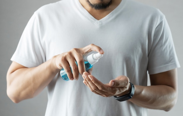 Asian bearded man in white shirt standing over grey background using rubbing alcohol portable spraying on his palms and hands