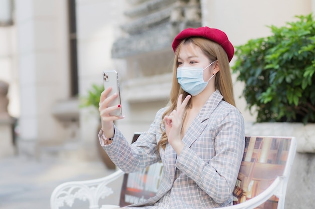 Asian beautiful female wears suit and red cap and holds smartphone for video call in her hands. She acts a two thumbs up sign showing victory while sits on bench in park outdoors while wearing a medic