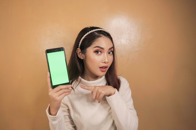 아시아의 아름다운 소녀는 흰색 셔츠와 함께 스마트폰에서 충격을 받은 녹색 화면을 보여줍니다