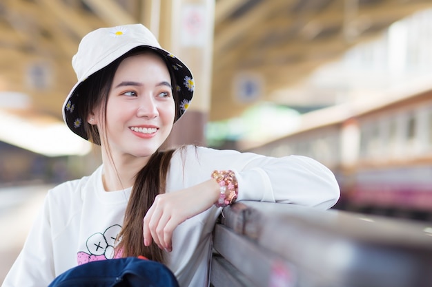 Asian beautiful girl in a longsleeved white shirt and a hat sits happy smilie in the train station