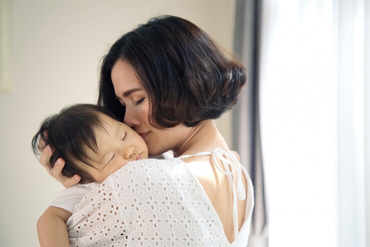 アジアの美しい母親が彼女の腕の中で眠っている赤ちゃんを抱いてと優しく子供にキスします。母親は赤ちゃんの頭を肩に乗せたまま目を閉じていました。愛と家族の関係のタッチ。