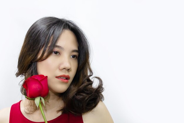 アジアの美しい女性は彼女の顔の横に赤いバラを持って目をそらしています。バレンタインデーと愛