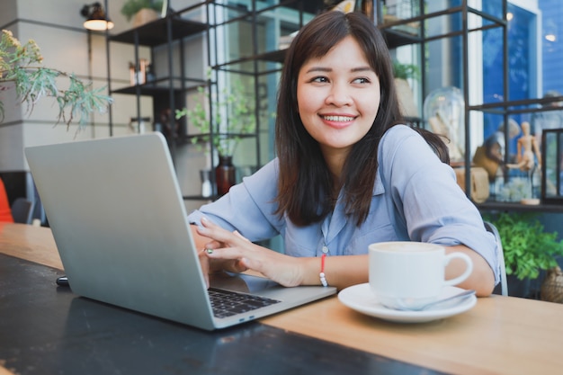 Bella donna asiatica in camicia blu facendo uso del computer portatile e bevendo caffè