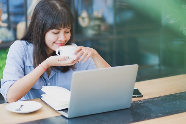 ラップトップを使用して、コーヒーを飲みながら青いシャツを着てアジアの美しい女性