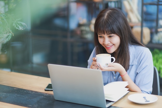 Bella donna asiatica in camicia blu facendo uso del computer portatile e bevendo caffè